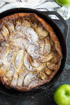 an apple pie with powdered sugar on top in a cast iron skillet next to two green apples