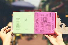 two hands holding an open book with pink and green pages in front of a cityscape