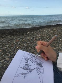 a person holding a pencil and drawing on paper next to the ocean with rocks in front of them