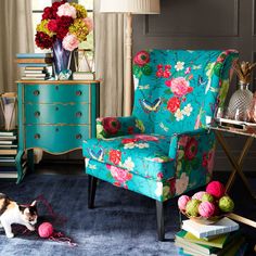 a cat sitting on the floor in front of a chair with flowers on it and a lamp