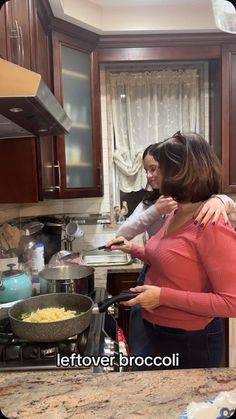 two women standing in a kitchen cooking food on the stove top and one is holding her arm around the other woman's shoulder