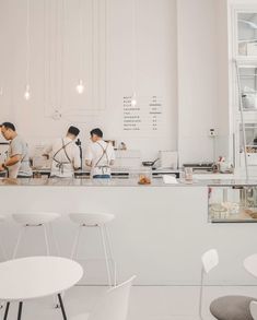 two men are preparing food in a white room with tables and chairs around them, while another man is working behind the counter