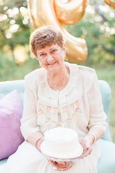 an older woman is holding a cake on a blue couch with balloons in the background