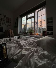 a laptop computer sitting on top of a bed in front of a window with the sun setting