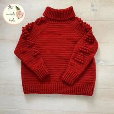 a red knitted sweater with pom poms on the collar and sleeves, sitting on top of a wooden floor