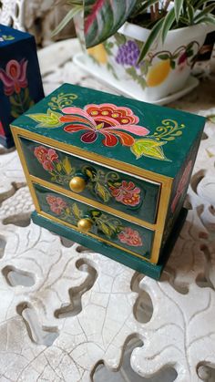 a green box with flowers painted on it sitting on a table next to a potted plant