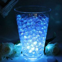 a glass filled with water sitting next to white roses and blue lights in the dark