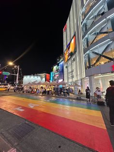 people are standing on the sidewalk in front of a large building at night with brightly colored floors