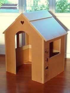 a cardboard dog house with the door open on a wooden floor in front of a window