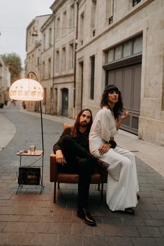 a man and woman sitting on a chair in the middle of an alleyway next to a lamp