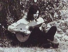 a man sitting on the ground playing a guitar in front of some trees and bushes