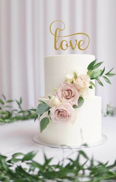 a white wedding cake with pink roses and greenery on top is decorated with the word love