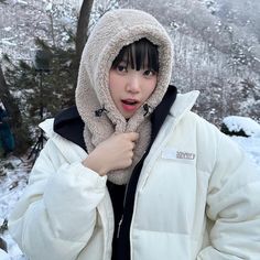 a woman wearing a white jacket and black hoodie standing in the snow with trees behind her