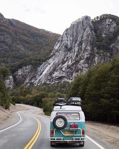 an old vw bus driving down the road in front of some mountains and trees