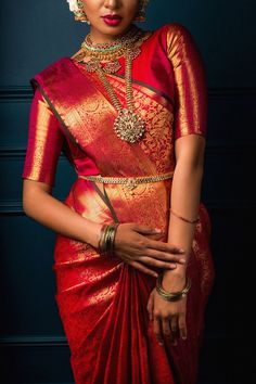 a woman in a red and gold sari with jewelry on her neck, standing against a