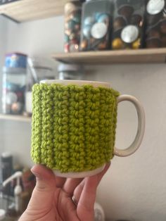 a hand holding a green crocheted coffee cup in front of a shelf with other items