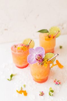 three glasses filled with drinks and garnished with flowers on the rim, sitting on a white surface