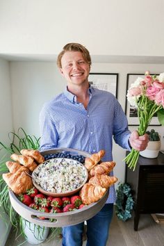 a man holding a platter full of food with flowers in the corner behind him