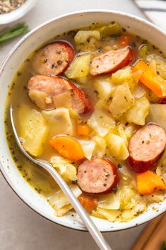 a bowl of soup with sausage, potatoes and carrots next to a wooden spoon