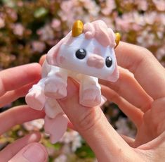 a hand holding a tiny toy cow with horns on it's head in front of pink flowers