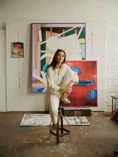 a woman sitting on top of a stool in front of a painting and holding a cat