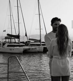 a man and woman standing next to each other on a boat in the water with sailboats behind them