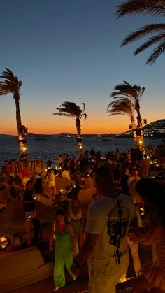 many people are sitting on the beach at night with palm trees in the foreground