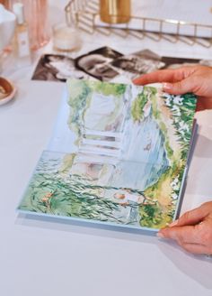 two hands holding an open book on top of a white table with other items in the background