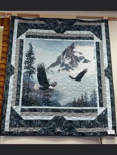 a quilt hanging on the wall with two birds flying in front of mountains and trees