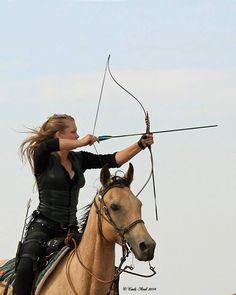 a woman in black riding on the back of a brown horse while holding an arrow