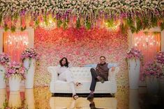 a man and woman sitting on a couch in front of a flower covered wall with pink flowers