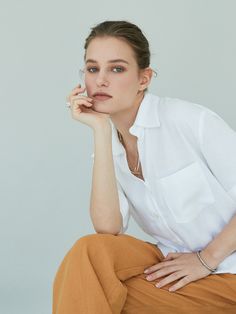 a woman sitting on the ground with her hand under her chin, wearing a white shirt and brown pants