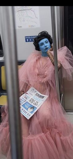 a woman with blue makeup sitting on a train reading a newspaper while wearing a pink dress