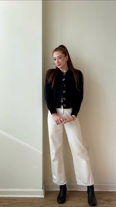 a woman standing in front of a wall wearing white pants and a black cardigan