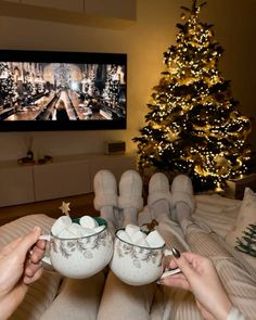 two people holding cups in front of a christmas tree with lights on the tv screen