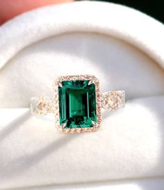 an emerald and diamond ring on top of a white cloth with a hand holding it