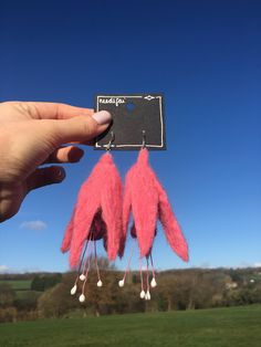 a pair of pink feather earrings hanging from the side of a green field with trees in the background