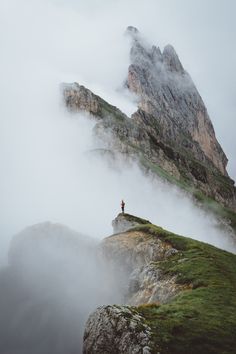 a person standing on top of a green hill next to a tall mountain covered in fog