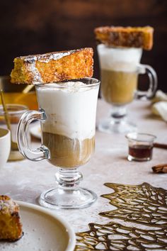 two glasses filled with coffee and cinnamon on top of a white table covered in food