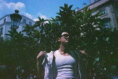 a man in white shirt and sunglasses standing next to green trees with buildings in the background