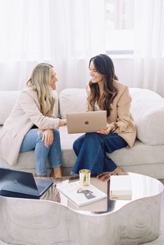 two women sitting on a couch talking to each other with laptops in front of them