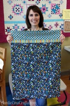 a woman holding up a piece of fabric in front of a quilted wall hanging