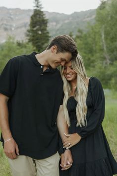 a man and woman standing next to each other in the grass with mountains behind them