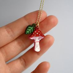 a hand holding a red and white mushroom necklace on a gold plated chain with green leaves