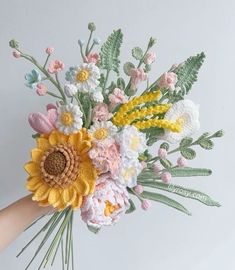 a hand holding a crocheted flower bouquet with yellow, white and pink flowers