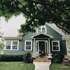 two people are sitting on the front steps of a green house with white trim and windows