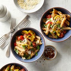 three bowls filled with pasta and vegetables on top of a white table next to silverware