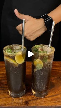 two glasses filled with ice and lemon on top of a wooden table next to a person