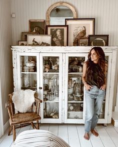 a woman standing in front of a china cabinet with pictures on the wall behind it