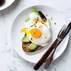 an egg and avocado toast on a white plate next to a knife and fork